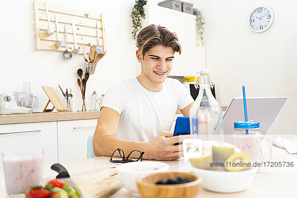 Lächelnder junger Mann  der ein Smartphone benutzt  während er am Tisch in der Küche sitzt