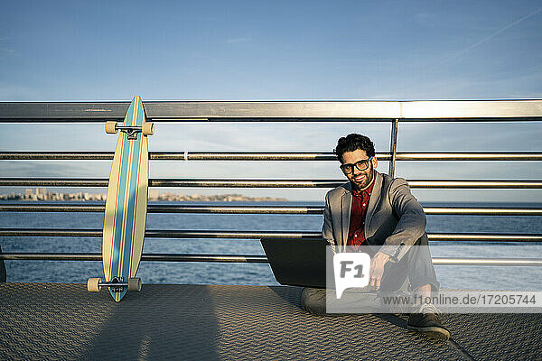 Lächelnder Geschäftsmann mit Laptop  der auf dem Geländer eines Piers sitzt