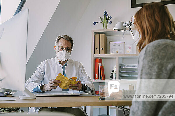 Doctor wearing protective face mask checking patient's vaccination certificate while sitting at office