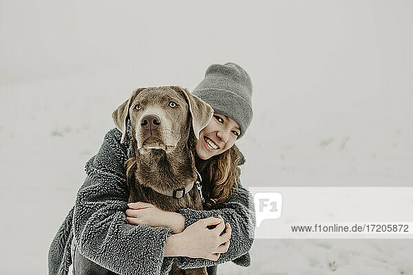 Porträt eines Mädchens im Teenageralter  das im Schnee hockt und einen Labrador Retriever umarmt