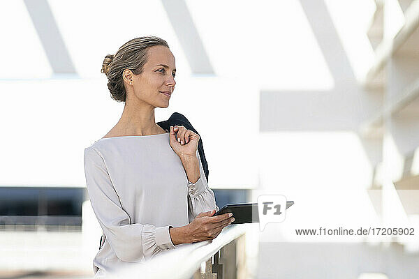 Geschäftsfrau mit digitalem Tablet und Jacke schaut weg  während sie auf einer Büroterrasse steht
