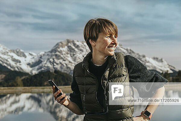 Lächelnde Frau mit Smartphone  die wegschaut  während sie am Prinzensee  Hochkönigmassiv  Salzburger Land  Österreich steht