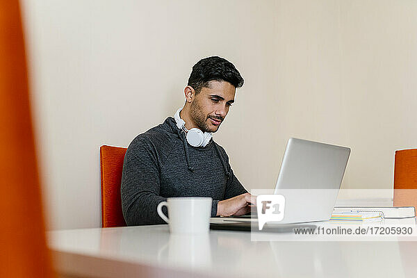 Junger Mann mit Kopfhörer  der einen Laptop benutzt  während er am Tisch im Wohnzimmer sitzt