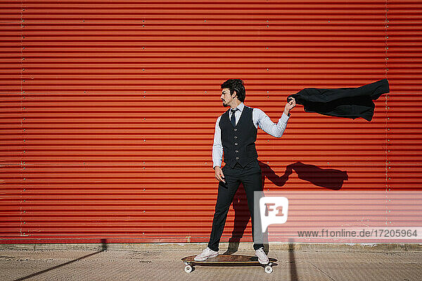 Männlicher Unternehmer hält Blazerjacke  während er auf einem Longboard an der Wand steht