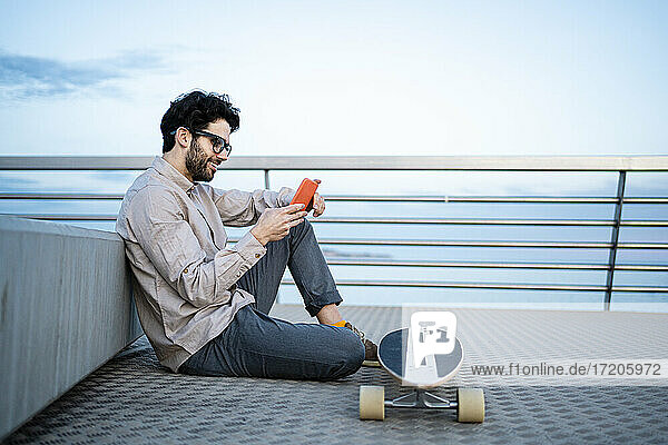 Lächelnder Geschäftsmann  der ein Mobiltelefon auf einem Longboard benutzt  während er auf einem Pier sitzt und sich gegen eine Bank lehnt
