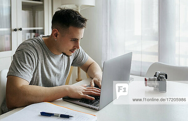 Junger männlicher Berufstätiger  der einen Laptop benutzt  während er ein Video mit einer Digitalkamera im Heimbüro aufnimmt