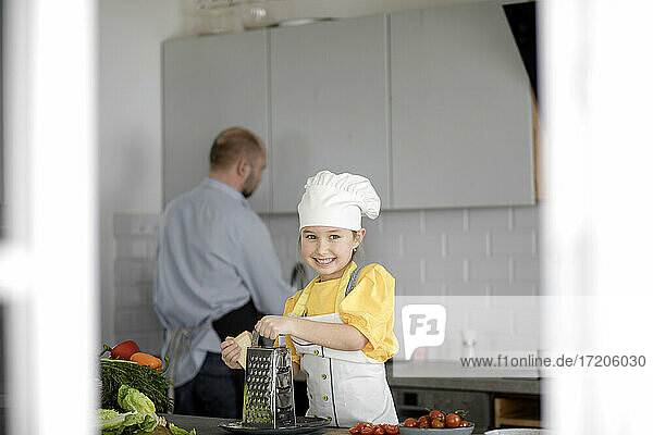 Lächelndes Mädchen mit Schürze und Kochmütze  das eine Reibe benutzt  während es mit seinem Vater im Hintergrund in der Küche steht
