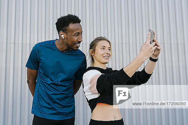 Female athlete with man taking selfie through mobile phone while standing against wall