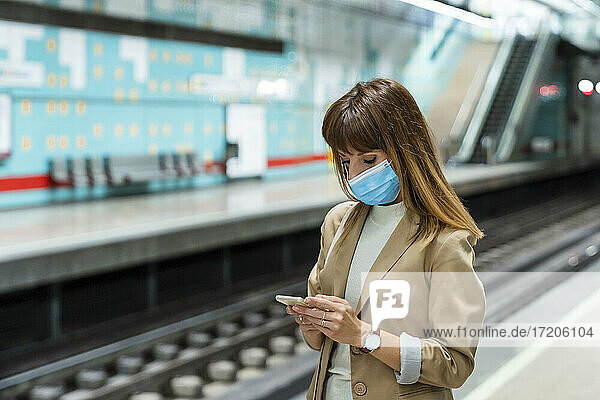 Woman with protective face mask using mobile phone while standing at subway station during pandemic