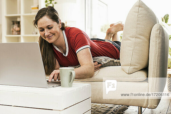 Mittlere erwachsene Frau  die einen Laptop benutzt  während sie auf dem Sofa im Wohnzimmer liegt