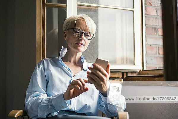 Reife Geschäftsfrau  die ein Mobiltelefon benutzt  während sie auf einem Stuhl am Fenster im Heimbüro sitzt