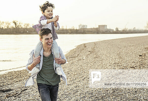 Cheerful young man walking while carrying boy on shoulders by lake