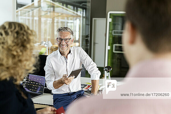 Smiling businessman with digital tablet discussing with colleagues at desk in office