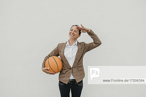Lächelnde weibliche Fachkraft mit Basketball  die vor einer weißen Wand steht und grüßt