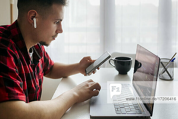 Männlicher Unternehmer mit In-Ear-Kopfhörern  der sein Smartphone in der Hand hält  während er im Heimbüro am Laptop arbeitet