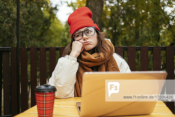 Gelangweilte Frau,  die mit ihrem Laptop in einem Straßencafé sitzt und wegschaut