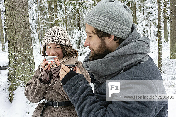 Couple drinking coffee while sitting in forest during winter