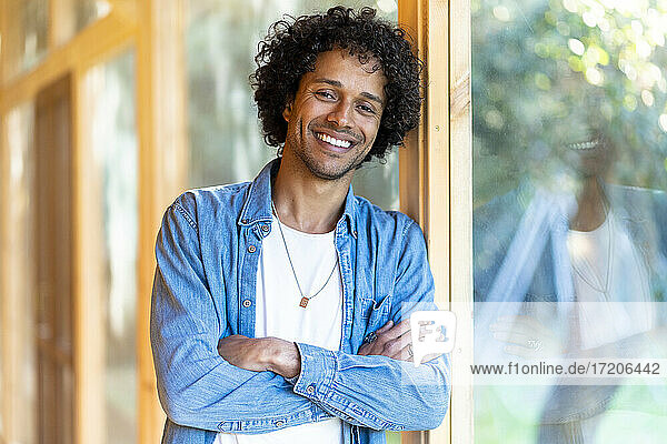 Lächelnder junger Mann  der sich in einem geräumigen Zimmer an ein Gartenfenster lehnt