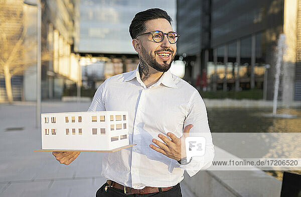 Lächelnder männlicher Architekt gestikuliert  während er ein Architekturmodell auf dem Fußweg hält