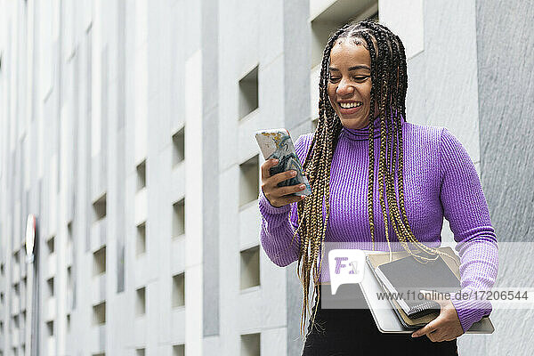 Lächelnde Frau mit Büchern  die ein Smartphone benutzt  während sie an einer grauen Wand steht