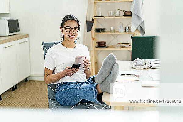 Junge Frau mit Kaffeetasse und Mobiltelefon  lächelnd zu Hause sitzend