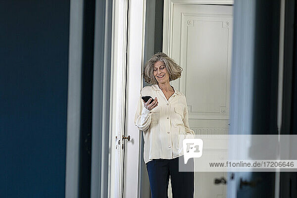 Lächelnde Frau  die ein Mobiltelefon benutzt  während sie zu Hause vor der Tür steht