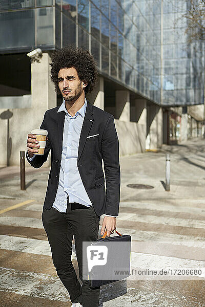 Geschäftsmann mit Laptoptasche und Kaffeetasse beim Überqueren der Straße an einem sonnigen Tag