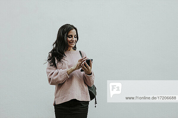 Lächelnde Frau  die ein Smartphone benutzt  während sie vor einer weißen Wand steht
