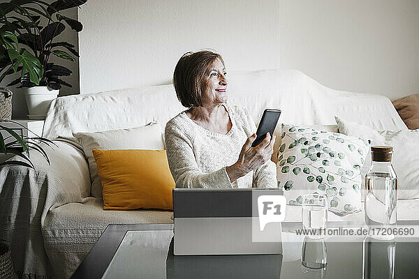 Smiling senior woman with laptop holding mobile phone while sitting on sofa at home