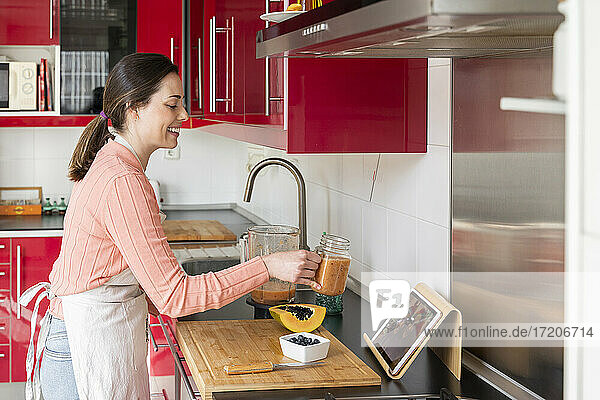 Lächelnde Frau  die Freunden bei einem Videoanruf ein Smoothie-Glas zeigt  während sie in der Küche zu Hause steht