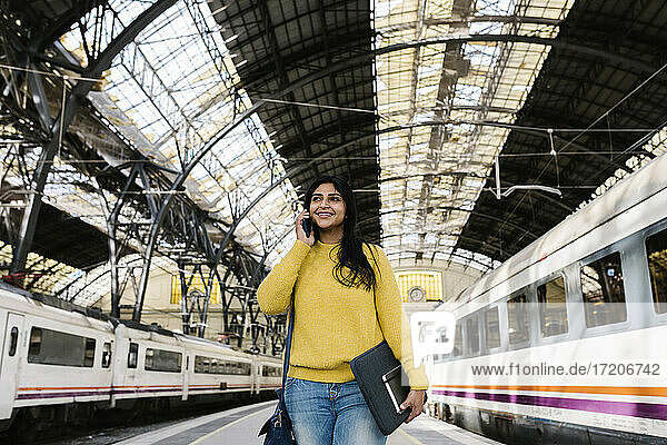 Lächelnde Frau mit Laptoptasche  die wegschaut  während sie auf dem Bahnsteig mit ihrem Smartphone telefoniert