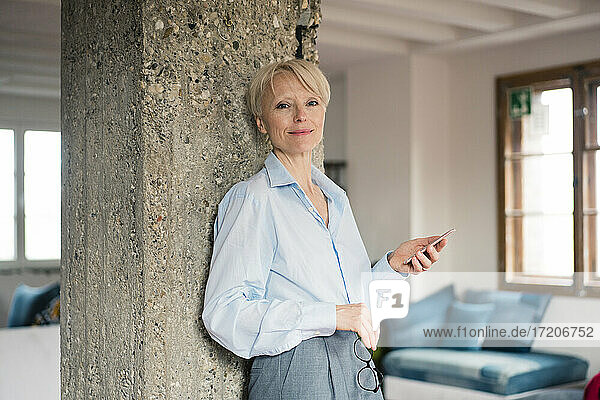 Smiling freelance worker holding mobile phone while leaning on column at home