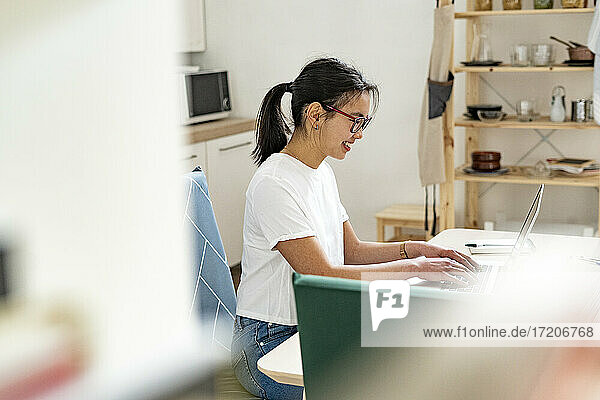 Smiling woman working on laptop while sitting in kitchen at home