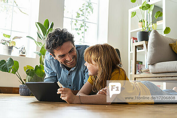 Lächelnde Tochter mit digitalem Tablet  die ihren Vater anschaut  während sie zu Hause im Wohnzimmer auf dem Boden liegt