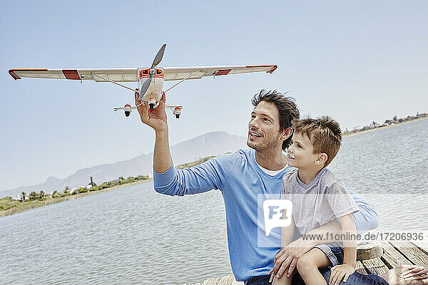 Vater hält Flugzeugspielzeug  während er mit seinem Sohn am See sitzt
