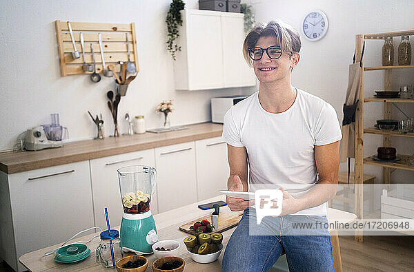 Lächelnder junger Mann mit digitalem Tablet auf dem Tisch in der Küche sitzend