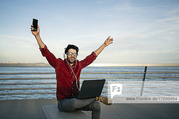 Sorgloser Geschäftsmann mit Laptop  der die Arme hebt  während er auf einer Bank am Meer über Kopfhörer Musik hört