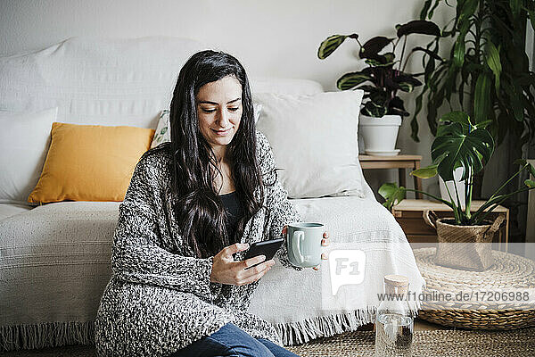 Lächelnde Frau  die eine Kaffeetasse hält  während sie ein Mobiltelefon auf dem Sofa im Wohnzimmer benutzt