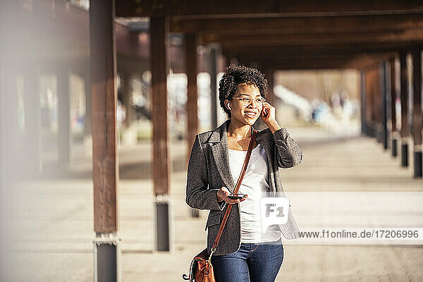 Smiling young woman with mobile listening music through in-ear headphones on sunny day