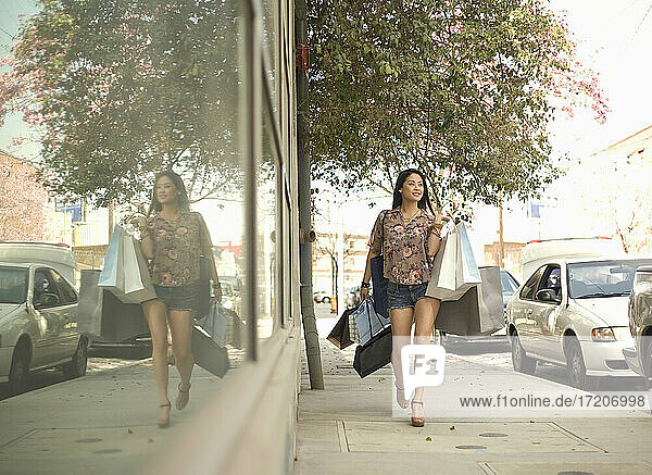 Junge Frau geht auf dem Bürgersteig und trägt Einkaufstaschen durch Glasfenster in der Stadt