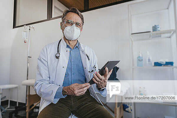 Männlicher Arzt hält ein digitales Tablet  während er in einer Klinik sitzt