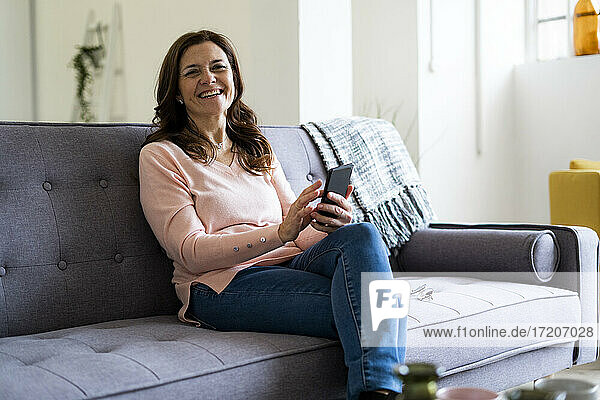 Glückliche reife Frau  die ein Mobiltelefon hält  während sie auf dem Sofa im Wohnzimmer sitzt