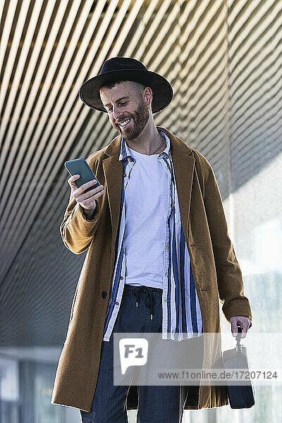 Mittlerer erwachsener Geschäftsmann mit Tasche  der im Freien stehend ein Mobiltelefon benutzt