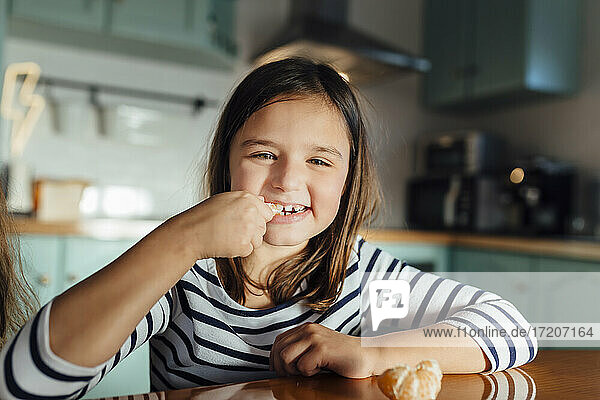Lächelndes Mädchen isst Mandarine  während es am Esstisch in der Küche sitzt