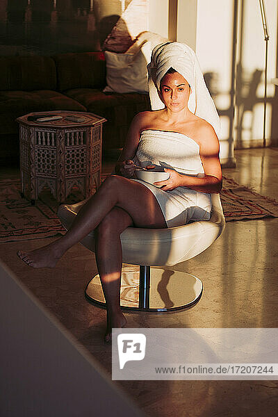 Entspannte Frau im Handtuch mit Obstschale  die wegschaut  während sie auf einem Stuhl zu Hause sitzt