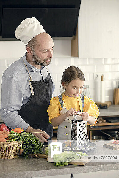Vater mit Schürze und Kochmütze bei der Essenszubereitung mit seiner Tochter zu Hause