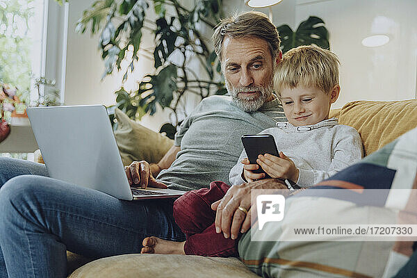 Father looking at boy using smart phone while sitting on sofa at home
