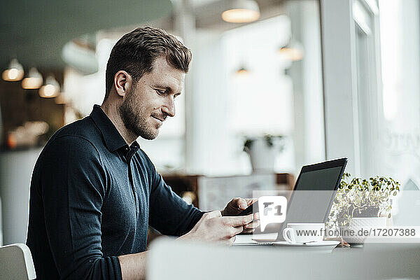 Mid adult businessman with laptop using smart phone while sitting at cafe