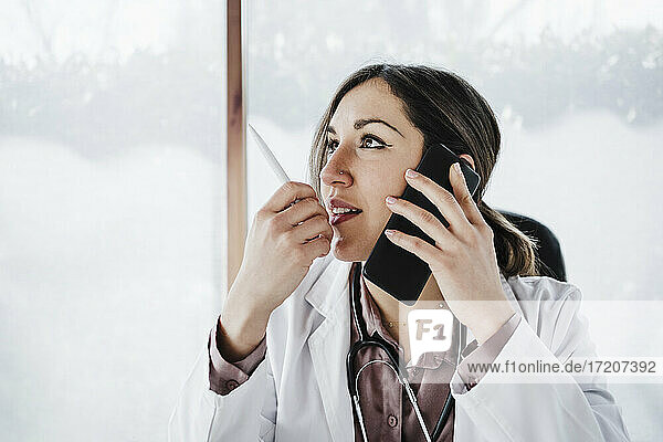 Nachdenkliche Ärztin  die wegschaut  während sie mit einem Mobiltelefon spricht