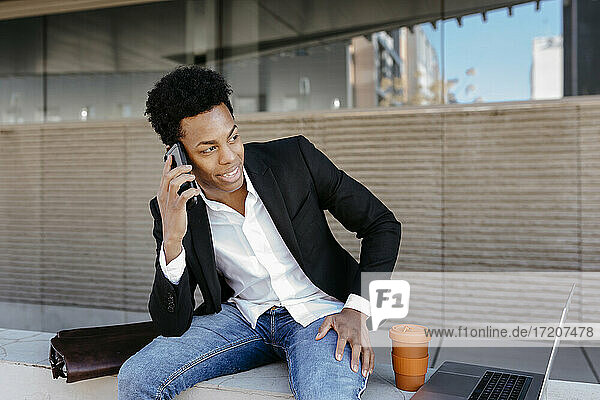 Lächelnder männlicher Geschäftsmann  der auf einer Stützmauer sitzend mit einem Smartphone spricht und dabei wegschaut